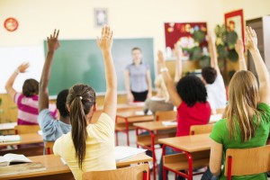 Group of teenagers sitting in classroom with raised hands.